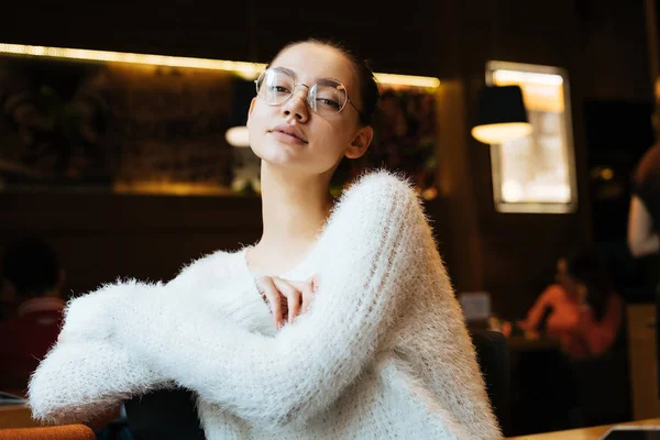 Серйозна молода дівчина фрілансер в білій куртці і окулярах сидить у затишному кафе і дивиться на камеру — стокове фото