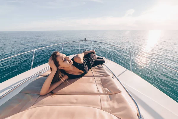 Роскошная стройная девушка в летней черной одежде лежит на своей белой яхте, наслаждаясь теплом и солнечным светом — стоковое фото