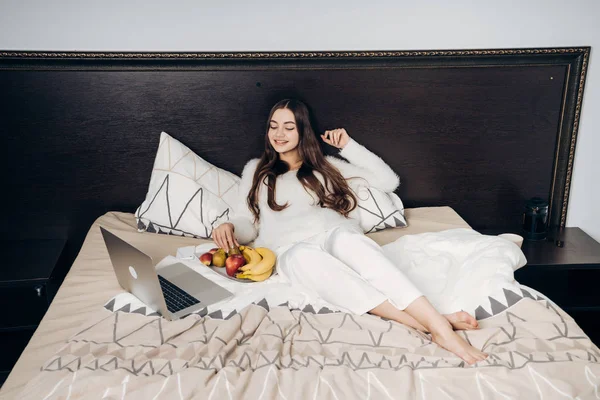 그녀의 노트북에 재미 있는 영화를 보이는 작업 후 침대에 젊은 여 자가 거짓말을 웃 고 과일을 먹는다 — 스톡 사진