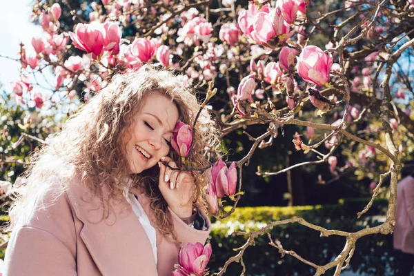 Mujer rizada feliz disfrutando del aroma de una magnolia floreciente en un parque al sol — Foto de Stock