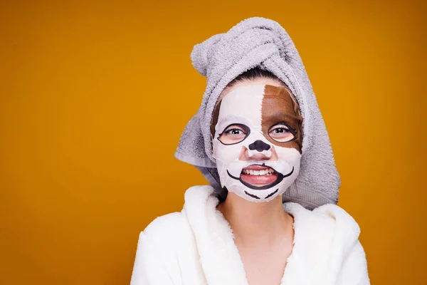 Rolig tjej med en handduk på huvudet och i en vit morgonrock efter dusch ler, på hennes ansikte en mask med en hunds ansikte — Stockfoto