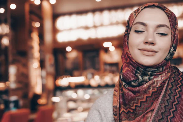 En trevlig Muslim tjej med huvudduk sitter i ett mysigt orientaliskt café, väntar på sin mat och leende — Stockfoto