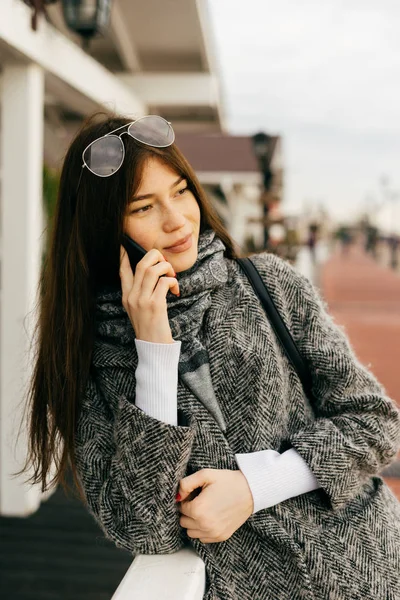 Belle fille de la ville aux cheveux bruns dans un manteau gris à la mode marchant dans les rues de la ville, parlant au téléphone, style de rue — Photo