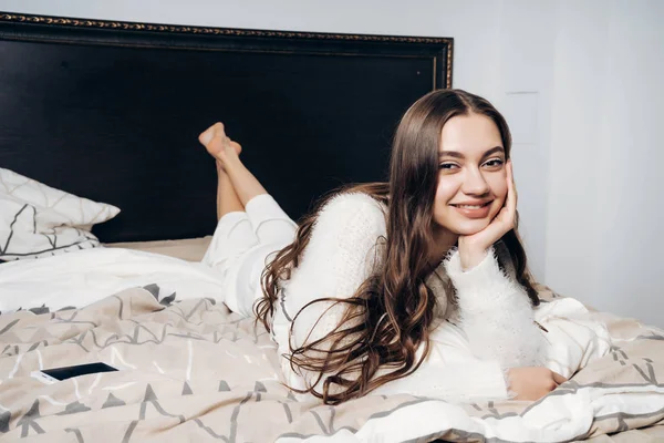 Счастливая длинноволосая девушка в пижаме лежит на кровати рано утром, в хорошем настроении, улыбаясь — стоковое фото