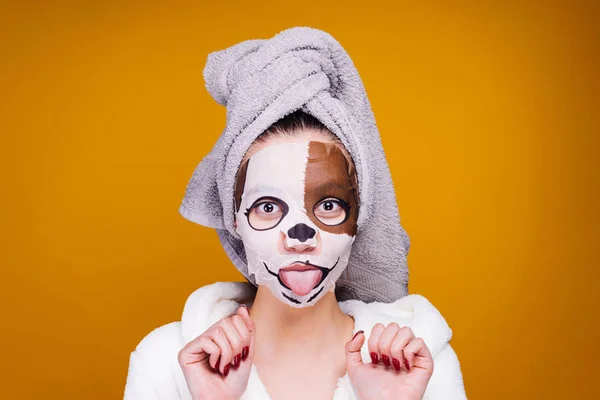 Grappig meisje met een handdoek op haar hoofd toont haar tong, op haar gezicht een nuttige hydraterende masker met een dog's gezicht — Stockfoto