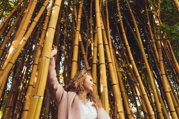 Curly stylish girl in a coat posing next to tall bamboo trees, enjoying the warmth — Stock Photo, Image