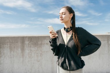 confident young girl in black sports clothes went to a workout in the open air, looks into her smartphone