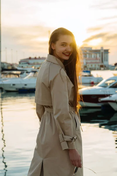 Uśmiechający się piękna, młoda dziewczyna z długimi włosami w beżowy płaszcz, stwarzające przez morze o zachodzie słońca — Zdjęcie stockowe