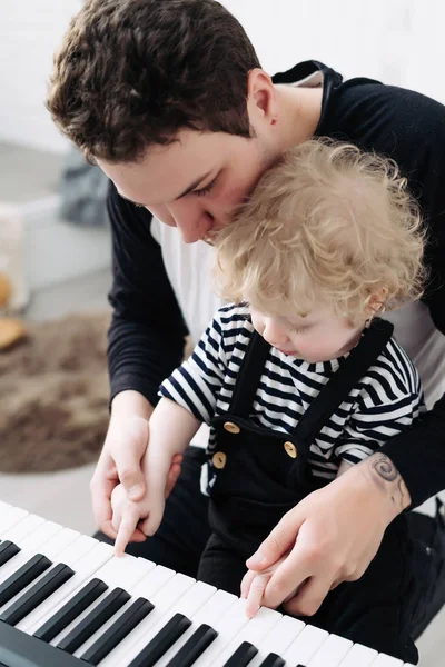 Premuroso giovane papà insegna al suo piccolo figlio riccio a suonare il pianoforte — Foto Stock