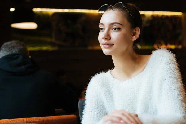 Красива молода дівчина-фрілансер в білому светрі сидить в кафе, посміхаючись і думаючи про роботу — стокове фото