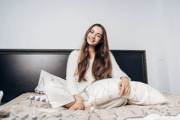 Gelukkig jong meisje met lang haar en in witte pyjama zit op het bed vroeg in de ochtend, heeft een goede slaap en glimlacht — Stockfoto