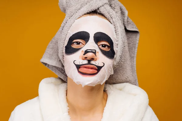 Buena chica con una toalla en la cabeza y en un albornoz después de una ducha, en su cara una máscara hidratante con una cara de panda — Foto de Stock