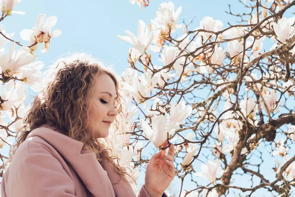 Elegante mujer rizada huele una magnolia floreciente en el parque, se regocija en primavera y calor — Foto de Stock