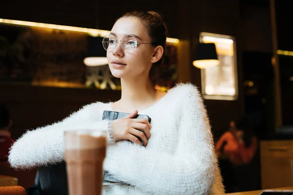 Красива молода дівчина студентка в білому піджаку і окулярах сидить в кафе після школи, дивлячись у вікно — стокове фото