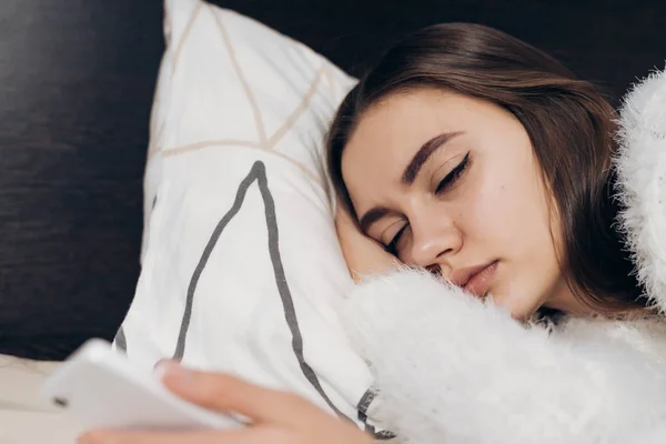 Süße schläfrige junge Mädchen im weißen Pyjama liegt in den frühen Morgenstunden im Bett, schaltet den Wecker auf ihrem Handy aus — Stockfoto