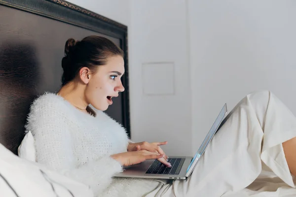 Joven sorprendida en pijama se sienta en la cama, descansando y viendo un interesante programa de televisión en su computadora portátil —  Fotos de Stock