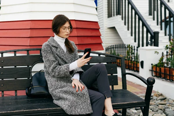 Élégante fille de la ville aux cheveux bruns portant des lunettes et un manteau gris assis sur un banc à l'extérieur, tenant un smartphone — Photo