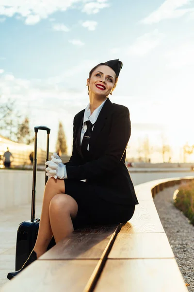 Улыбается красивая стюардесса девушка в форме сидит в парке с чемоданом, смотрит в небо — стоковое фото