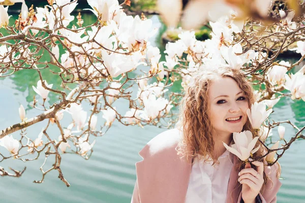 Smiling curly woman in pink coat posing in park next to blooming magnolia — Stock Photo, Image