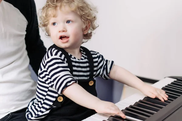 Kudrnatá malý chlapeček s modrýma očima hraje na klavír, sedí na klíně s jeho otcem — Stock fotografie
