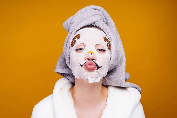 Grappig meisje met een handdoek op haar hoofd en in een badjas na een regenbui, op haar gezicht een hydraterende masker — Stockfoto