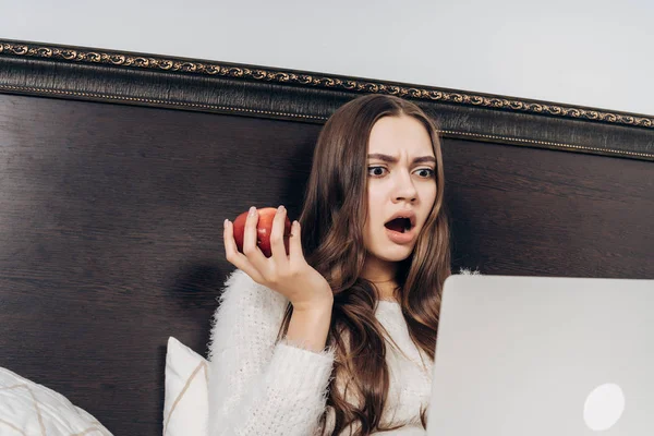 Сердитая удивленная девушка смотрит интересный фильм на ноутбуке, сидит в постели и ест яблоко — стоковое фото
