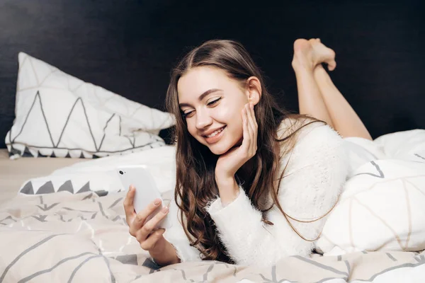 Счастливая улыбающаяся девушка в пижаме лежит на кровати, глядя в свой смартфон, хорошее настроение — стоковое фото