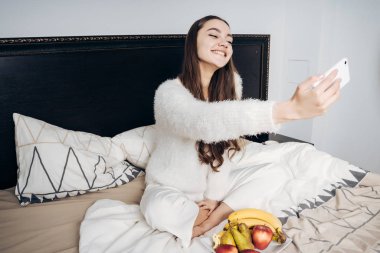 gülümseyen ve selfie onun Smartphone'da yapma sevimli uykulu kız yatakta sabah erken, oturan pijama