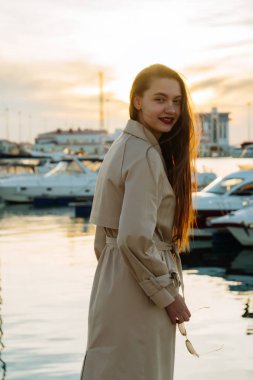 deniz kenarında günbatımında poz ve gülümseyerek, onun yat için bekleyen trendy ceket güzel genç kadın