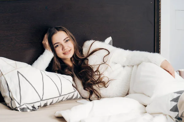 Schattig jong meisje in witte pyjama ligt in bed vroeg in de ochtend, heeft een goede slaap en glimlacht — Stockfoto