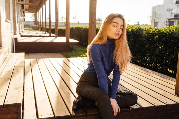 Fiduciosa ragazza bionda in maglione blu siede su una panchina di legno al sole nel parco — Foto Stock