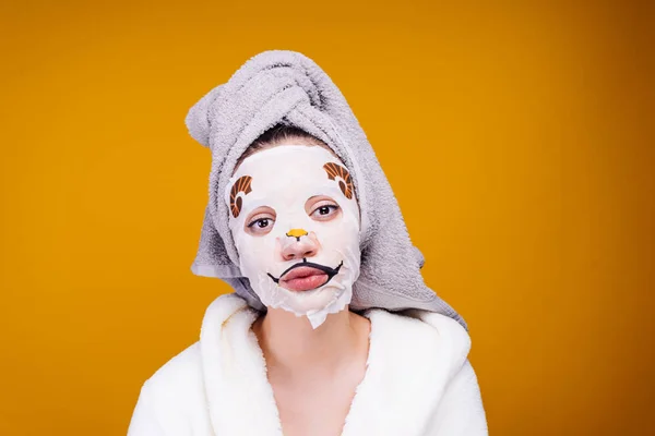 Een jong meisje met een handdoek op haar hoofd en een badjas in witte kleren zorgt voor zichzelf, op haar gezicht een hydraterende masker — Stockfoto