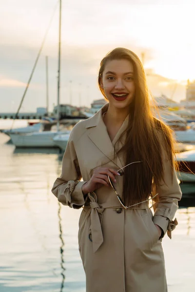 Feliz de cabelos longos bela menina segurando óculos de pé no porto ao pôr do sol e sorrindo — Fotografia de Stock