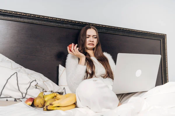 Menina de cabelos longos em pijama senta-se na cama à noite, come frutas úteis e assiste filme de terror em seu laptop — Fotografia de Stock