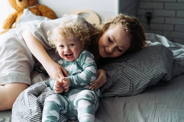 Familia feliz, madre joven jugando con su pequeño hijo rizado en la cama, divirtiéndose — Foto de Stock