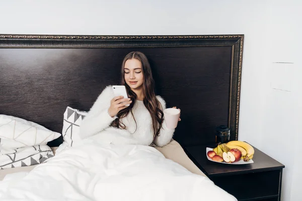 Hermosa mujer joven descansando en la cama después de un duro día de trabajo, mirando en su teléfono inteligente — Foto de Stock