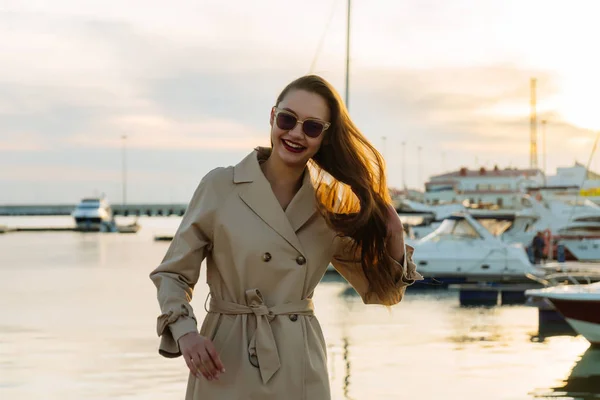 Atrakcyjny dziewczynka długowłosy w okularach słonecznych i beżowy płaszcz jest stojący w porcie morskim w godzinach wieczornych i uśmiechając się — Zdjęcie stockowe