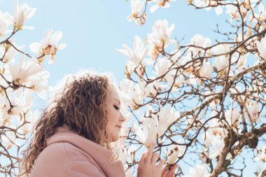 kıvırcık genç kadın mavi gökyüzü altında çiçek açan bir Manolya koklama pembe bir ceket