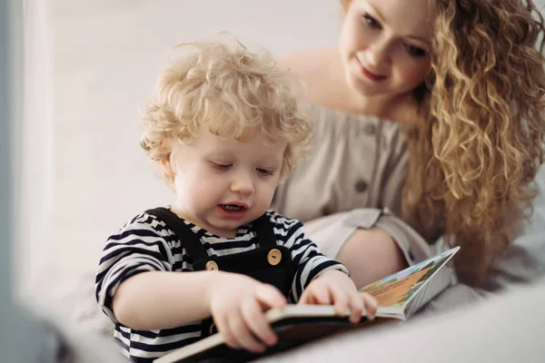 A caring curly mother plays with her little child a boy, read a book together — Stock Photo, Image