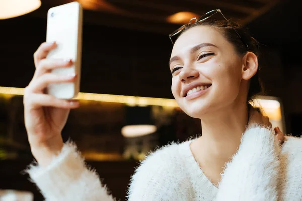 Szczęśliwy uśmiechający się student dziewczyna w białym swetrze siedzi w kawiarni, wygląda na swoim smartfonie, zapisuje komunikat — Zdjęcie stockowe