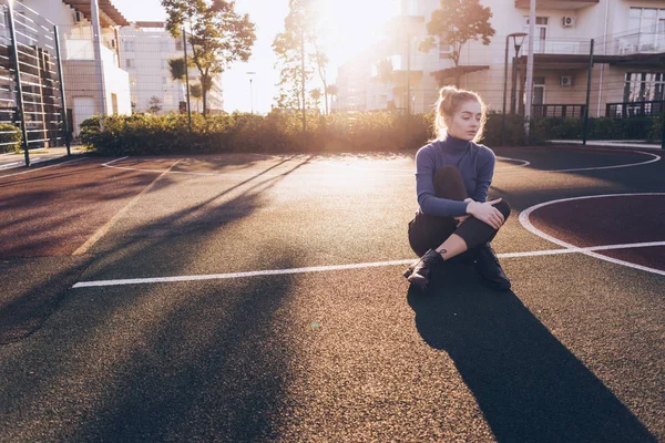 Stijlvolle blond model meisje in blauwe trui zit op de grond in een gebied van de sport buiten in de zon — Stockfoto