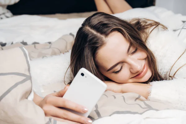 Милая улыбающаяся девушка в пижаме лежит в постели рано утром, держит смартфон и не хочет вставать — стоковое фото