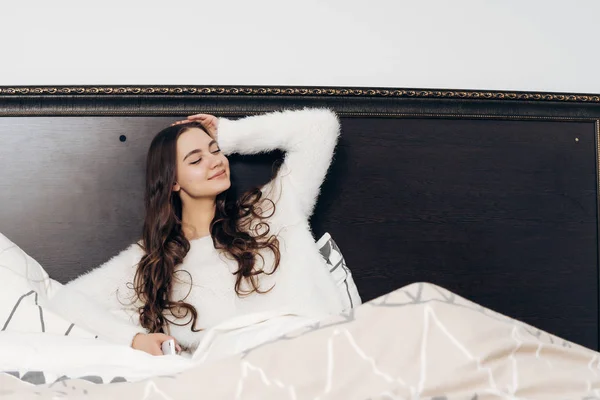 Χαριτωμένο υπνηλία μακρυμάλλης κορίτσι σε λευκό πιτζάμες έγκειται στο κρεβάτι νωρίς το πρωί και δεν θέλετε να σηκωθεί, χαμόγελα — Φωτογραφία Αρχείου