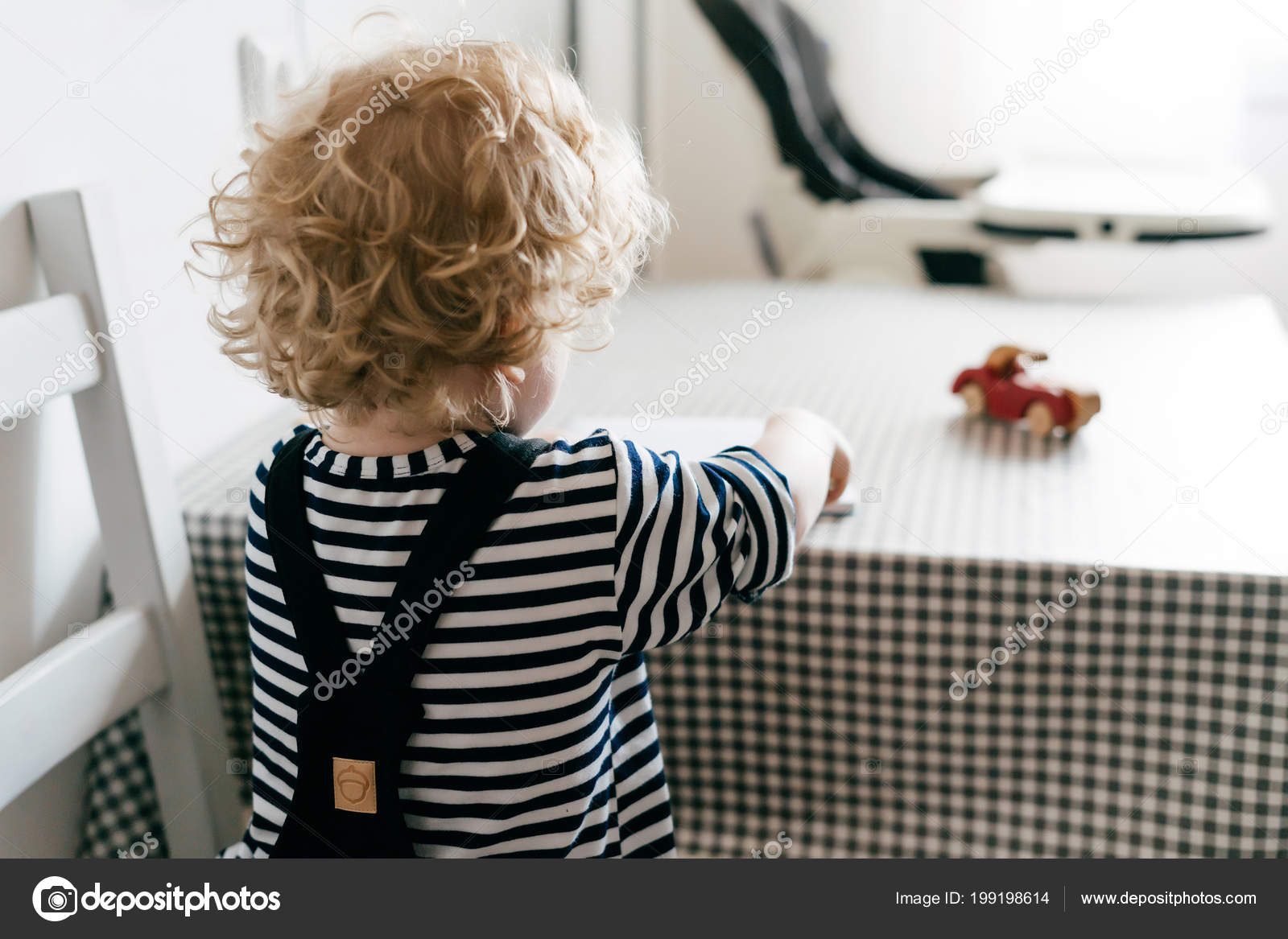 Curly Little Baby Boy In Overalls Sitting At Table In Kitchen And
