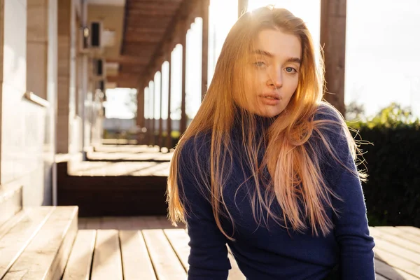 Confident beautiful blonde girl in blue trendy sweater sits on a bench in the park in the sun — Stock Photo, Image