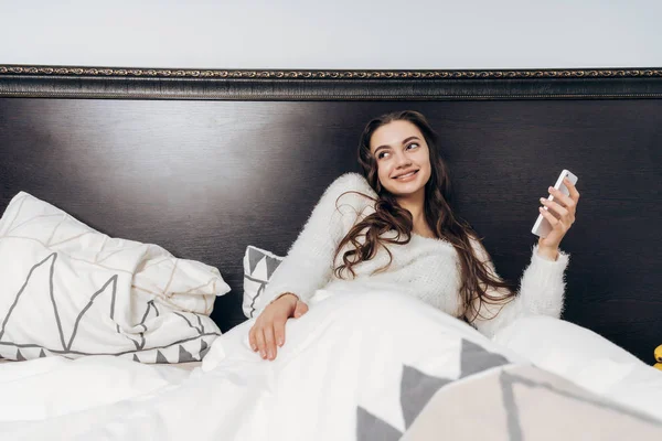 Alegre joven en pijama se encuentra en la cama temprano en la mañana, sostiene el teléfono inteligente y sonríe — Foto de Stock