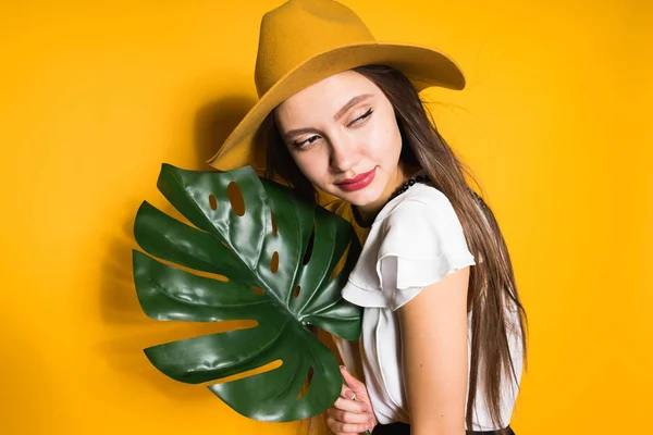 유행 모자에 자신감 긴 머리 소녀 모델 보유 녹색 잎과 오렌지 배경에 포즈 — 스톡 사진