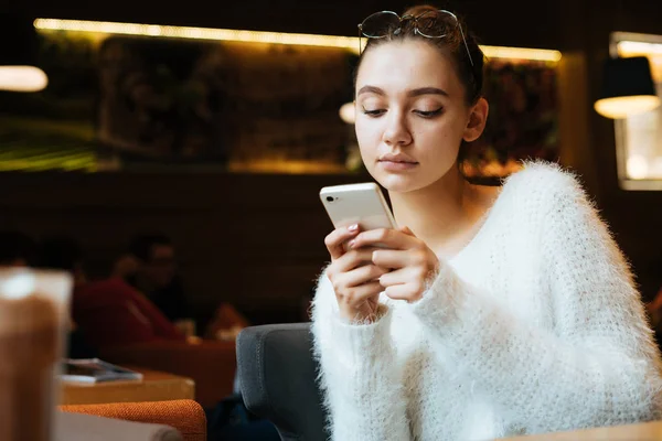 A thoughtful young girl freelancer in a white sweater sits in a cafe, writes a message on her smartphone — Stock Photo, Image
