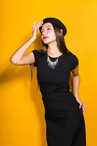 Luxurious confident girl in a black trendy dress and hat posing against a yellow wall background — Stock Photo, Image