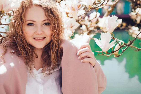 Elegant curly woman in a pink coat posing against a pond background, smiling, enjoying the sun and spring — Stock Photo, Image
